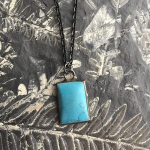 May include: A rectangular turquoise pendant necklace with a dark chain. The pendant is a vibrant blue-green color, set in a silver-toned metal frame. The chain is a dark, possibly black, color. The necklace is displayed on a textured, gray background.