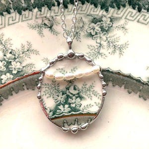 May include: A silver necklace with a pendant made from a vintage porcelain plate with a green floral pattern. The pendant is surrounded by a silver beaded border and features a row of white pearls.