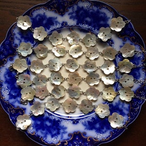 Puede incluir: Colección de botones con forma de flor hechos de nácar iridiscente, conectados por cadenas de color plateado. Los botones están dispuestos sobre un plato con estampado azul y blanco, creando una estética delicada y vintage.