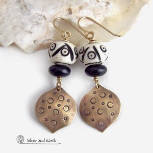 May include: Handmade earrings featuring cream-colored beads with black geometric designs, black beads, and bronze-colored leaf-shaped pendants with circular patterns. The earrings have gold-colored ear wires. The earrings are from Silver and Earth.