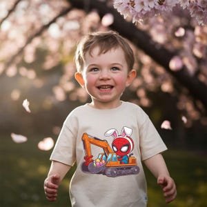 Könnte beinhalten: Ein kleines Kind trägt ein beiges T-Shirt mit einem Cartoon-Bild von Spiderman, der Hasenohren trägt und einen mit Ostereiern gefüllten Bagger fährt.