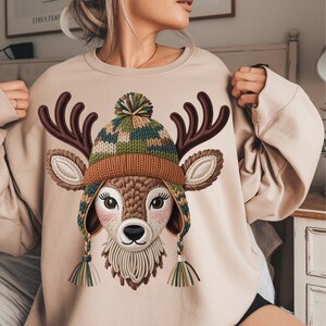 Könnte beinhalten: Beiges Sweatshirt mit einer Illustration eines Hirsches mit Wintermütze. Der Hirsch hat braune Geweihe und eine Strickmütze mit grünen, braunen und blauen Mustern. Das Sweatshirt ist neutralfarben und für Freizeitkleidung geeignet.