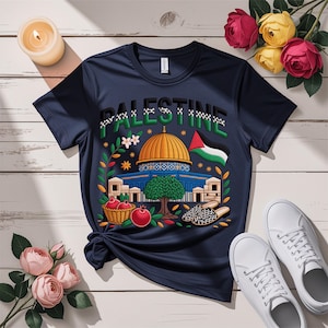 Könnte beinhalten: Marineblaues T-Shirt mit dem Wort "PALESTINE" und einer Illustration des Felsendom, einer palästinensischen Flagge und anderen kulturellen Elementen. Weiße Turnschuhe liegen neben dem Shirt.