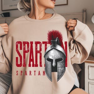 Peut inclure: Sweat-shirt beige avec un motif "SPARTAN" rouge vieilli et une illustration détaillée d'un casque spartiate. Le mot "SPARTAN" est imprimé en dessous du casque. Le casque a un plumet rouge.