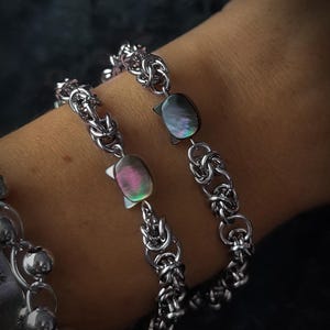 May include: Silver-toned bracelets with intricate chain links and iridescent oval accents. One bracelet features a crescent moon and star design. The image is set against a dark background, highlighting the jewelry's details.