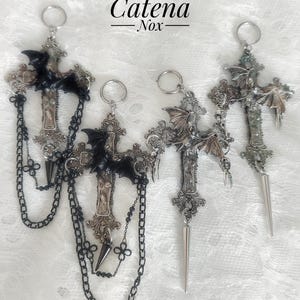 May include: Four metal cross pendants with the text Catena Nox at the top. Two pendants feature black bat figures and black chains, while two feature silver bat figures and silver spikes. Each cross has intricate detailing and a circular ring loop.