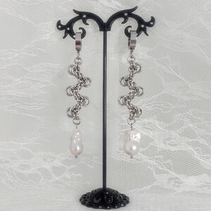 May include: A pair of silver-toned chainmail earrings featuring a dangling white baroque pearl at the base of each link structure. The earrings are displayed on a black stand with the text Catena Nox printed in the bottom left corner.