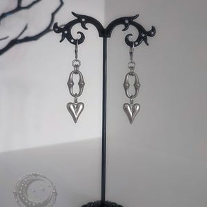 May include: A pair of silver-toned drop earrings with heart-shaped pendants. The earrings are displayed on a black metal stand with a decorative base. The earrings have a chain-like design and are suspended from a hook.