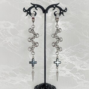 May include: A pair of silver-toned chainmail earrings hanging from a black display stand. Each earring features a series of interlocking metal rings, a dark grey cross-shaped charm, and a pointed metal spike at the bottom. The text Catena Nox is visible.
