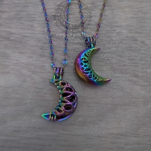 Könnte beinhalten: Zwei Halsketten mit schillernden Mond-Anhängern. Die Anhänger haben ein detailliertes Design und sind an passenden Ketten befestigt. Die Farben wechseln von Blau über Grün zu Lila. Die Halsketten werden auf einer hellgrauen Holzoberfläche präsentiert.