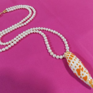 May include: A white pearl necklace with a long, orange and white seashell pendant. The necklace is on a pink background.