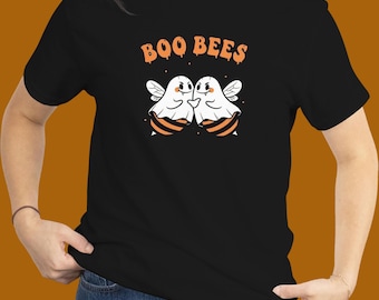 May include: Black t-shirt featuring the text "BOO BEES" in orange above a graphic of two ghost-like bees with white wings and orange and black stripes. The bees are embracing. The shirt is worn by a person.