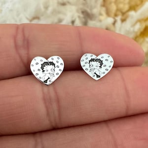 Puede incluir: Un par de pendientes de plata en forma de corazón con una imagen en blanco y negro de Betty Boop dando un beso. Los pendientes están rodeados de un patrón de pequeños corazones.