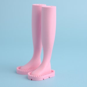 Puede incluir: Un par de piernas de muñeca rosas con una superficie texturizada. Las piernas están unidas a una base con un borde festoneado.