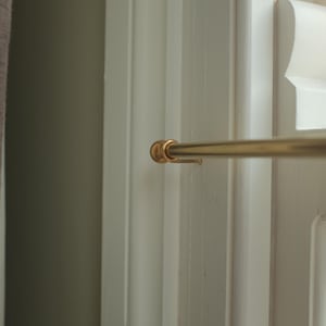 May include: A gold metal curtain rod with a round finial on the end. The rod is mounted to a white wall.
