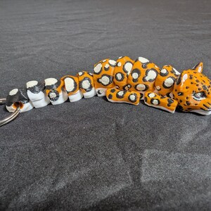 May include: A segmented, articulated keychain shaped like a jaguar, with black, white, and orange spots. The keychain has a silver metal ring.