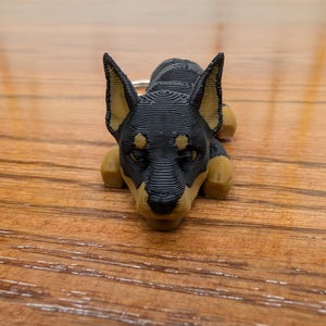 May include: A black and tan 3D printed dog keyring. The dog is lying down with its head resting on its paws.
