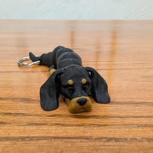 May include: A black and brown dachshund dog keychain. The dog is lying down with its head resting on its paws. The keychain has a silver metal ring.