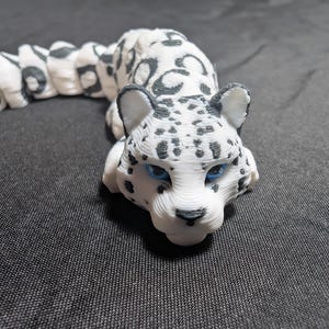 Peut inclure: Un porte-clés léopard blanc et gris avec des yeux bleus. Le léopard est couché, avec une chaîne de perles blanches et grises attachée à un porte-clés argenté. Le porte-clés est un article imprimé en 3D.