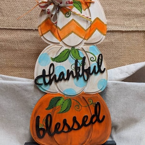 May include: A stack of three painted wooden pumpkins with the words "thankful" and "blessed" on them. The top pumpkin has a chevron pattern in orange and white, the middle pumpkin has blue polka dots, and the bottom pumpkin is orange. The pumpkins are on a black wooden stand.