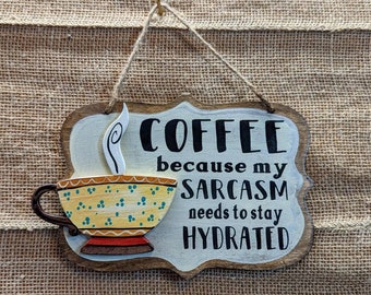 May include: A wooden sign with a white background and black text that reads "Coffee because my sarcasm needs to stay hydrated". A yellow and blue coffee cup with a steam curl is painted on the sign.