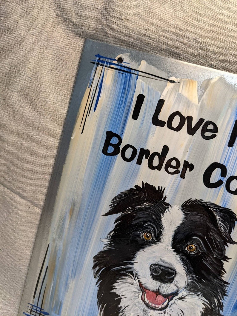 Puede incluir: Un letrero pintado con un perro Border Collie con pelaje blanco y negro y ojos marrones. El letrero tiene las palabras "I Love Border Collies" en letras negras sobre un fondo azul y blanco.