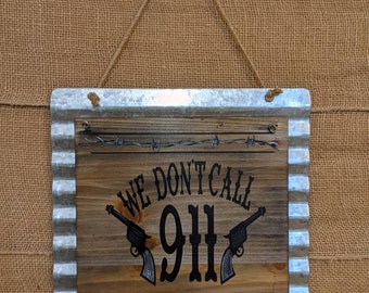 May include: A rustic metal sign with a wood background. The sign says "We Don't Call 911" with two black pistols on either side of the number 911. The sign is surrounded by a barbed wire design.