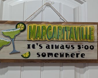 May include: Wooden sign with the word "MARGARITAVILLE" in green and yellow, and the phrase "It's always 5:00 somewhere." Two margarita glasses and a lime slice are also featured. The sign is hanging by a rope.