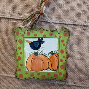 May include: A green and orange framed wooden sign with a black bird perched on a pumpkin. The sign has a polka dot pattern and the bird is looking to the left.