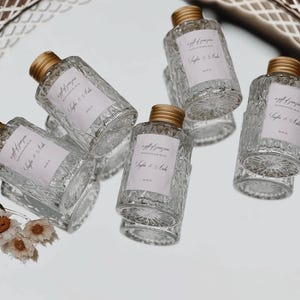 May include: Five clear glass bottles with gold lids and labels that say "a gift of joy from our wedding day" with the names "Sophia & Andre" on each label. The bottles are on a white surface.