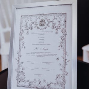 Op de afbeelding: Een zilver ingelijst document met ingewikkelde bloemenranden en Arabische kalligrafie bovenaan. De tekst bevat de namen Nur & Engin, datums en lijnen voor handtekeningen met de labels Gelin, Damat, Sahit en Imam, gedrukt op wit papier.