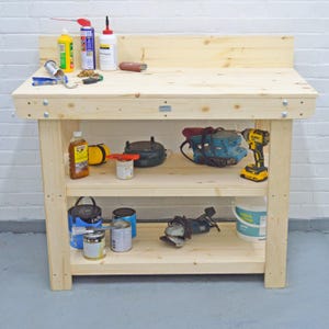 May include: A wooden workbench with two shelves, featuring a variety of tools and supplies, including paint cans, a power drill, a grinder, and a bottle of wood stain.