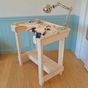 May include: A wooden workbench with a vice, tools, and a lamp. The workbench is made of natural wood and has a light brown finish. The workbench is standing on a wooden floor.