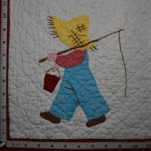 May include: A white quilted fabric square featuring an appliqué of a scarecrow figure wearing a yellow hat, red and white checkered shirt, and blue overalls. The figure carries a brown fishing pole and a red bucket. A ruler shows a 13-inch width.