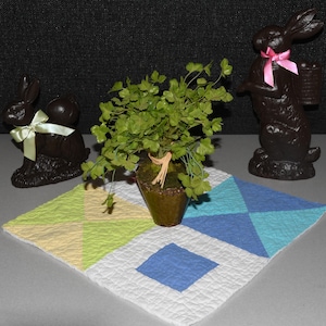 May include: A green potted plant with a brown pot sits on a white, blue, and yellow patchwork quilt. Two chocolate bunny figurines are on either side of the plant. One bunny has a green ribbon and the other has a pink ribbon.