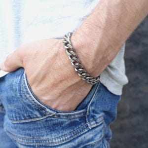Puede incluir: Una pulsera de cadena de eslabones de color plateado. La pulsera está hecha de eslabones gruesos e interconectados. La pulsera se lleva en la muñeca, con la persona que lleva una camiseta gris y vaqueros azules.
