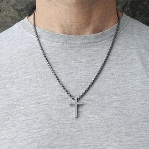 Op de afbeelding: Een zilverkleurige kruisvormige hangerketting. Het kruis heeft een eenvoudig ontwerp en hangt aan een ketting. De ketting wordt gedragen op een grijs T-shirt.