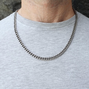 Puede incluir: Un collar de cadena de eslabones de color plateado. El collar está hecho de eslabones entrelazados y se lleva alrededor del cuello. La persona lleva una camiseta gris.