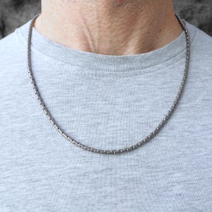 Peut inclure: Un collier argenté au design texturé. Le collier est porté autour du cou, reposant sur un t-shirt gris. La chaîne est composée de petits maillons rectangulaires reliés. Un accessoire simple et élégant.
