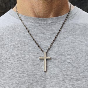Könnte beinhalten: Ein silberfarbener Kreuzanhänger hängt an einer Kette. Das Kreuz hat ein schlichtes, minimalistisches Design. Die Halskette wird über einem grauen T-Shirt getragen.