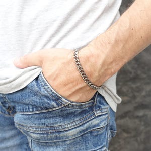 Op de afbeelding: Een zilveren gourmet armband wordt om een pols gedragen. De armband is gemaakt van in elkaar grijpende schakels. De persoon draagt een grijs t-shirt en een blauwe spijkerbroek. De armband is een stijlvol accessoire.