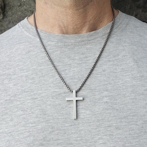 May include: A silver-colored cross pendant necklace. The cross is a simple, minimalist design. The chain is a dark, textured metal. The necklace is worn against a gray t-shirt.
