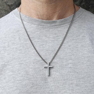 Op de afbeelding: Een zilverkleurige kruisvormige hangerketting. Het kruis heeft een eenvoudig, minimalistisch ontwerp en hangt aan een bijpassende ketting. De ketting wordt gedragen op een grijs T-shirt.