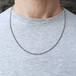 Könnte beinhalten: Eine silberfarbene Panzerketten-Halskette. Die Kette wird vor einem grauen T-Shirt gezeigt. Die Halskette ist ein schlichtes, klassisches Design, das für den Alltag geeignet ist. Die Kette ist etwa 50 cm lang.