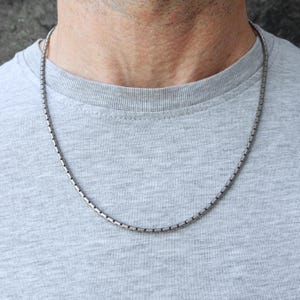 Peut inclure: Un collier en chaîne à maillons rectangulaires de couleur argentée. Le collier est drapé autour du cou d'une personne, reposant sur un t-shirt gris. La chaîne est composée de petits maillons rectangulaires.