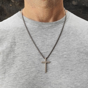 May include: A silver cross pendant necklace. The cross is a simple, classic design, suspended from a dark chain. The necklace is worn against a gray t-shirt.