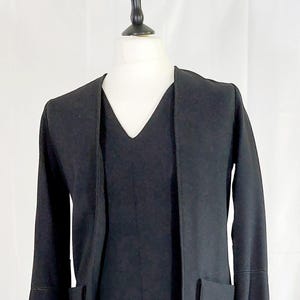 Op de afbeelding: Een zwart tweedelig ensemble op een mannequin. De outfit bestaat uit een knielange jurk met een V-hals en een bijpassend jasje met driekwart mouwen en twee voorzakken. De jurk en het jasje zijn gemaakt van een effen zwarte stof.