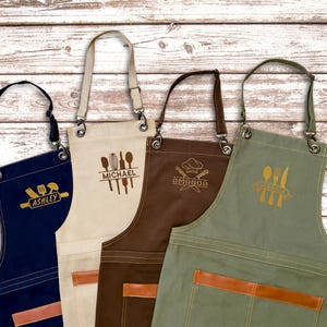 May include: Four personalized aprons in navy, cream, brown, and olive green, each with a unique kitchen-themed design and name. The aprons have leather accents and adjustable straps, perfect for cooking and baking.