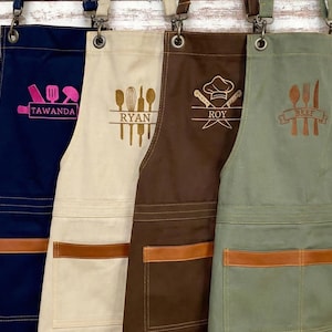 May include: Four aprons in navy, beige, brown, and olive green, each with a different design and name. The navy apron has pink kitchen tools and the name "TAWANDA". The beige apron has kitchen tools and the name "RYAN". The brown apron has a chef's hat and knives with the name "ROY". The olive green apron has a spoon, fork, and knife with the word "BEEF".