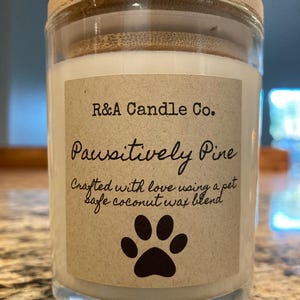 Peut inclure: Un bocal en verre transparent pour bougie avec un couvercle en bois. L'étiquette indique "R&A Candle Co. Paussitively Pine Crafted with love using a pet safe coconut wax blend". Une empreinte de patte noire est centrée sur l'étiquette.